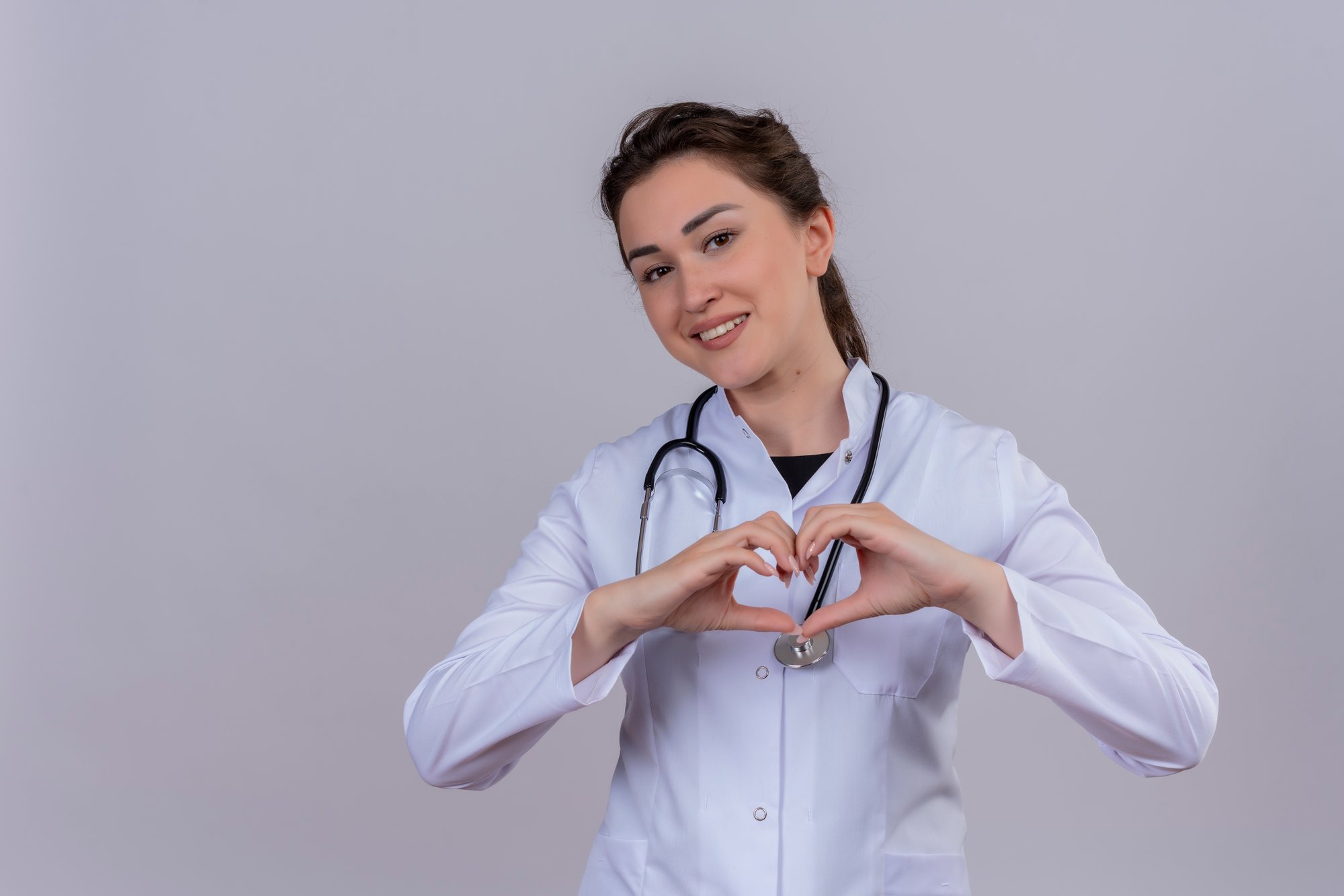 smiling-young-doctor-wearing-medical-gown-wearing-stethoscope-shows-heart-gesture-white-wall